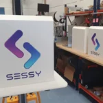 Thumbnail of  Witte Sessy thuisbatterij met paars-blauw logo in een werkplaats, met meerdere units op een houten werkbank.