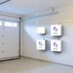 Thumbnail of  Drie witte Sessy thuisbatterijen en een omvormer gemonteerd aan een grijze garagemuur naast een roldeur.