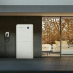 Thumbnail of  Wit Sigenstor Battery 8.0 kWh systeem met LED-indicator, geplaatst naast een buitenmuur met zicht op een huiskamer.